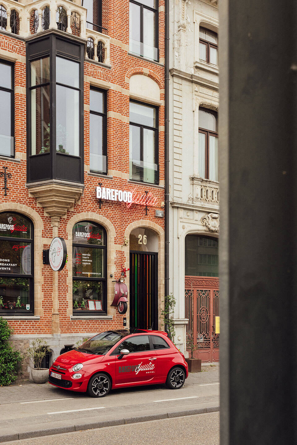 Vorderansicht des BareFoodGiulia Hotels in einem restaurierten Herrenhaus mit roter Ziegelfassade, großen Fenstern und einem roten Fiat 500 mit Hotellogo davor geparkt.